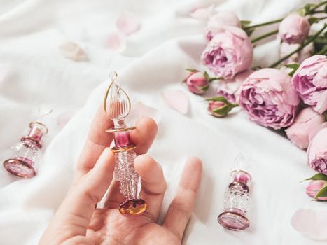 Three graceful bottles for perfume or essential oil on white crumpled fabric. Woman holds pink glass bottle with eastern ornament. Pink rose bouquet and petals as decoration.