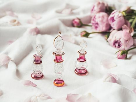 Three graceful bottles for perfume or essential oil on white crumpled fabric. Pink glass bottles with eastern ornament. Pink rose bouquet and petals as decoration.