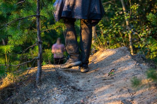 hiking man legs with shoes and gray pants somewhere in summer forest, close-up with selective focus. Blurred girl silhouette in the background.