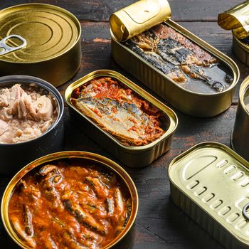 Tin cans for fish with different types of seafood, opened and closed cans with Saury, mackerel, sprats, sardines, pilchard, squid, tuna, over dark wood old table close up side ciew.