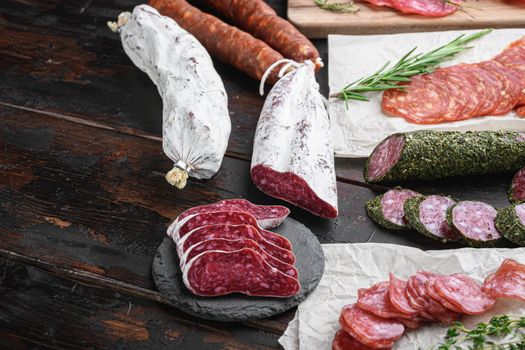 Variety of dry cured chorizo, fuet and other sausages cut in slices with herbs on dark wooden background.