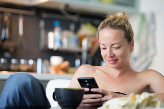 Young smiling cheerful woman indoors at home kitchen using social media on phone for video chatting and staying connected with her loved ones. Stay at home, social distancing lifestyle.