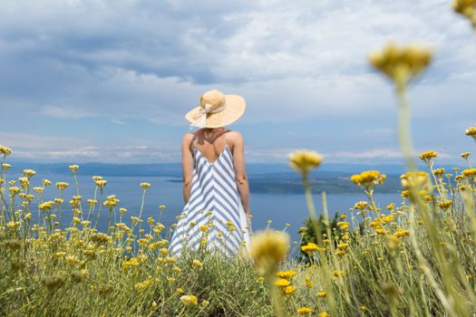 Rear view of young woman wearing striped summer dress and straw hat standing in super bloom of wildflowers, relaxing while enjoing beautiful view of Adriatic sea nature, Croatia. Focus on flowers.