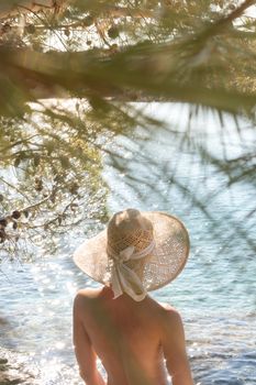 Rear view of topless beautiful woman wearing nothing but straw sun hat realaxing on wild coast of Adriatic sea on a beach in shade of pine tree. Relaxed healthy lifestyle concept.