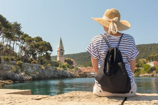 Woman traveler wearing straw summer hat and backpack, sittingat edge of stone pier, enjoying beautiful panoramic view of Veli Losinj, Losinj island, Croatia.