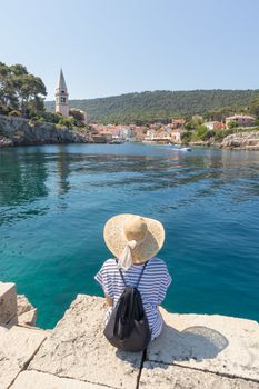 Woman traveler wearing straw summer hat and backpack, sittingat edge of stone pier, enjoying beautiful panoramic view of Veli Losinj, Losinj island, Croatia.