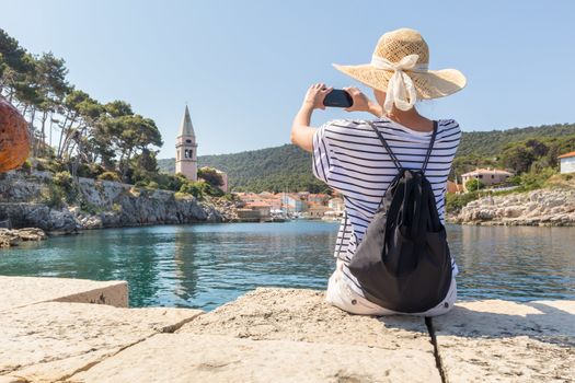 Woman traveler wearing straw summer hat and backpack, sittingat edge of stone pier, taking photo of beautiful panoramic view of Veli Losinj, Losinj island, Croatia.