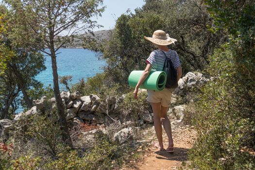 Young active feamle tourist wearing small backpack and yoga mat walking on coastal path among pine trees looking for remote cove to swim alone on seaside in Croatia. Travel and adventure concept.