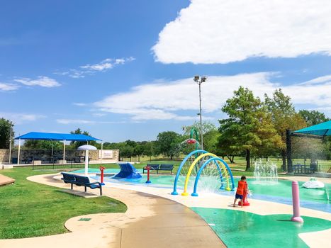 Rear view Asian toddler boy playing at splash park near Dallas, Texas, America. Colorful recreation site with splashing water fountains for kids summertime activities