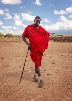 Unknown village near Amboselli park, Kenya - April 02, 2015: Unknown Masai warrior posing for tourists in traditional bright red robe, leaning on his stick.