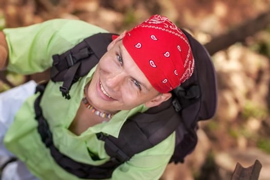 View from top on young man with green shirt and red bandana looking up and smiling, wearing backpack.
