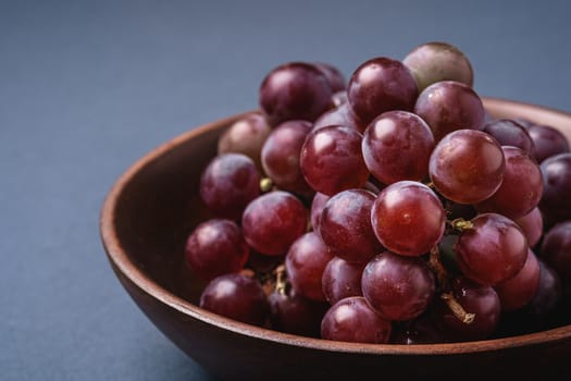 Fresh ripe grape berries in brown wooden bowl on blue grey minimal background, angle view macro