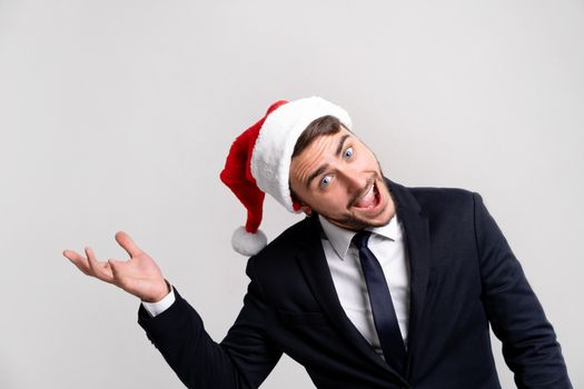 Young handsome caucasian guy in business suit and Santa hats on white background in studio smilie and showing hand to the side. Close up portrait business person with Christmas mood Holiday banner