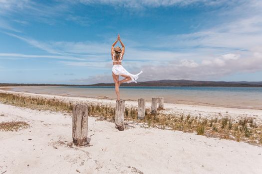 Beautiful woman balances elegantly on rustic timber post by water in yoga pose wearing swimwear and sheer sarong flowing in wind.
She is in an idyllic location in summer.