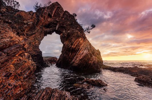 Sunrise sky at the towering pyramidal rock arch formation on the jagged coast line, Striped veins texture on the rock and its arch acts as a window to the ocean