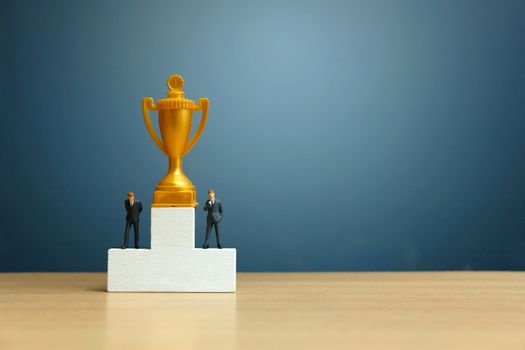 Miniature business concept - businessman standing in front of white winner podium with golden trophy