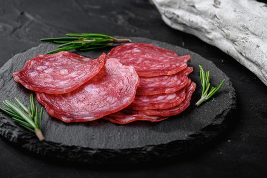Dry cured, whole and slices cuts of salchichon sausage on black textured surface.