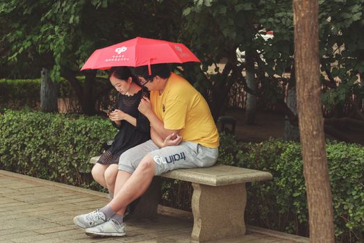 Harbin, Heilongjiang, China - September 2018: Couple sitting on bench. Man and woman under umbrella