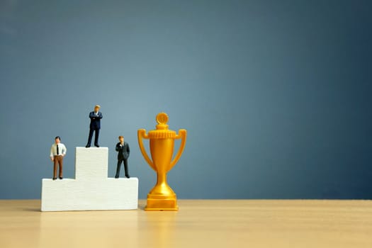 Miniature business concept - businessman standing in front of white winner podium with golden trophy