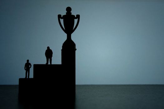 Business strategy conceptual photo - Silhouette of miniature businessman standing on podium to reach winning trophy