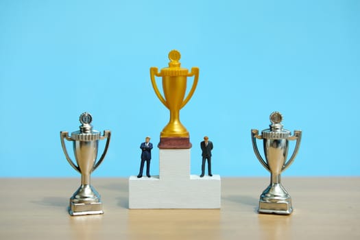 Miniature business concept - businessman standing on white winner podium with golden and silver trophy