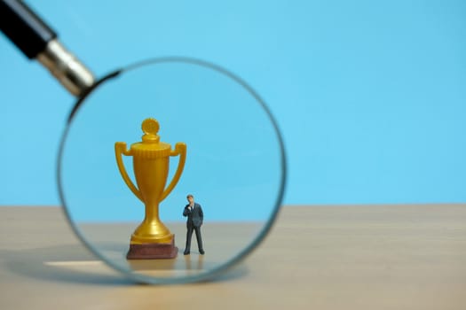 Miniature business concept - businessman standing in front of golden trophy and magnifier glass