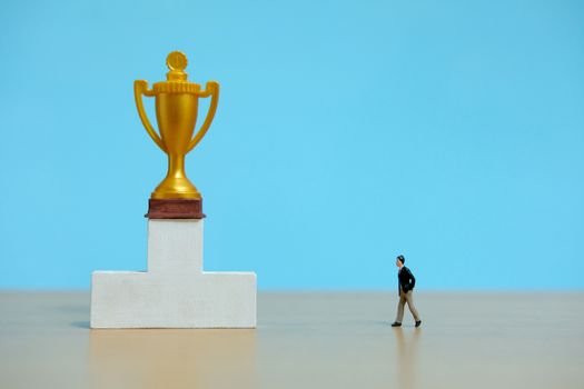 Miniature business concept - businessman walking toward golden trophy above white podium