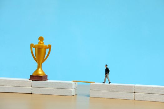 Miniature business concept - businessman walking toward golden trophy above white bridge