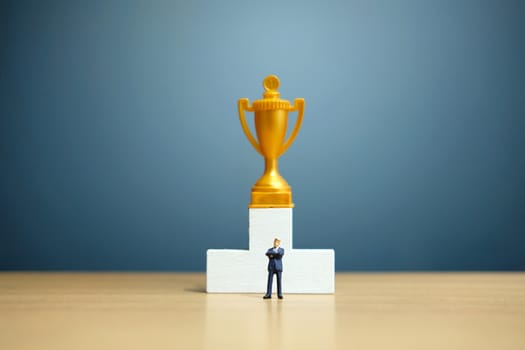 Miniature business concept - businessman standing in front of white winner podium with golden trophy