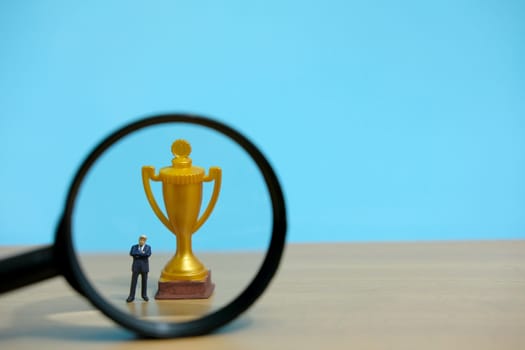 Miniature business concept - businessman standing in front of golden trophy and magnifier glass