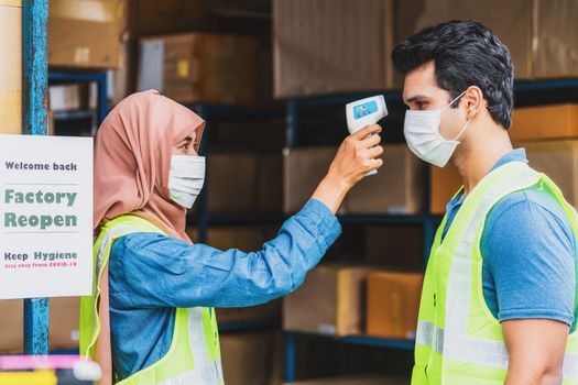 Muslim woman worker uses Medical Digital Infrared Thermometer measure temperature to Indian man worker with safety clothes before start to work after warehouse reopen again, new normal concept
