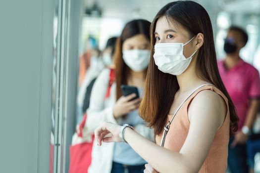 Young Asian woman passenger wearing surgical mask and checking the time when waiting bus at station with crowned people when traveling in big city at Covid outbreak, Infection and Pandemic concept