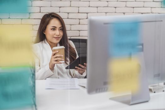 Asian Business woman holding paper coffee cup and mobile phone when working with technology computer over the the post it on the mirror at office,startup and entrepreneur,workshop and strategy concept