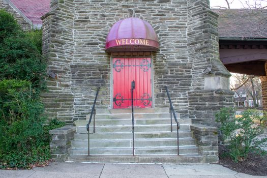 A Bright Red Door in a Cobblestone Wall With a Welcome Sign Above and Steps Leading Up to It and a Bush on the Side