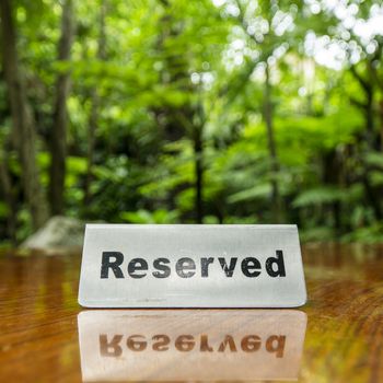 Reserved sign made out stainless steel plate on a laminated wooden table of a restaurant with trees and forest in the background.