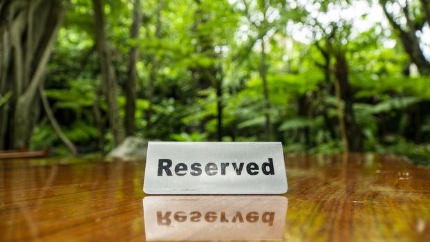 Reserved sign made out stainless steel plate on a laminated wooden table of a restaurant with trees and forest in the background.