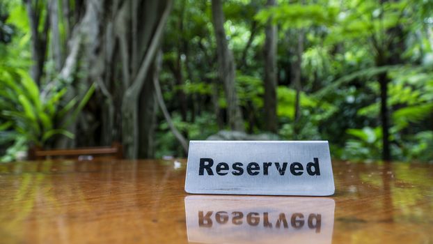 Reserved sign made out stainless steel plate on a laminated wooden table of a restaurant with trees and forest in the background.