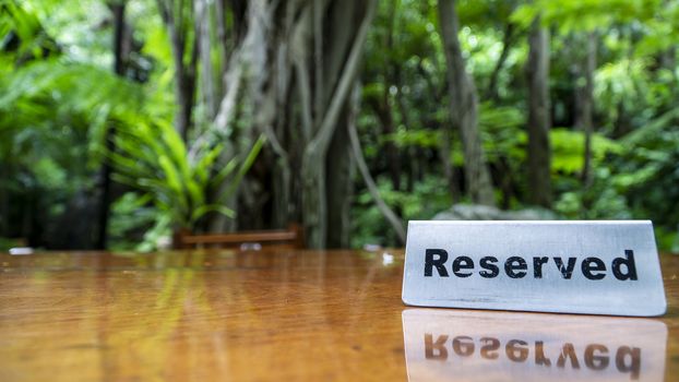 Reserved sign made out stainless steel plate on a laminated wooden table of a restaurant with trees and forest in the background.