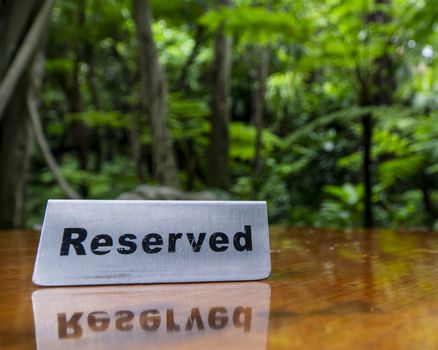 Reserved sign made out stainless steel plate on a laminated wooden table of a restaurant with trees and forest in the background.