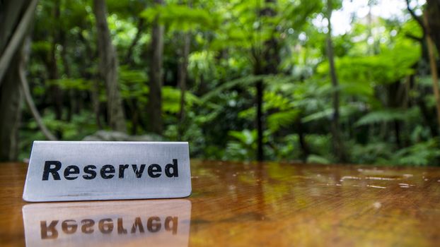 Reserved sign made out stainless steel plate on a laminated wooden table of a restaurant with trees and forest in the background.