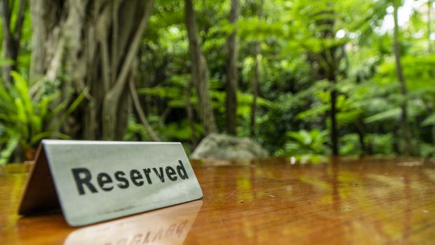 Reserved sign made out stainless steel plate on a laminated wooden table of a restaurant with trees and forest in the background.