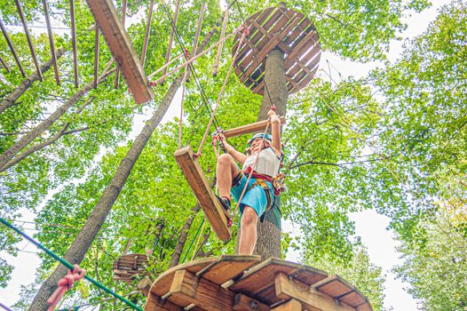 a young boy in outfit and a helmet holds on to the ropes near the round wooden platform in Forest adventure park
