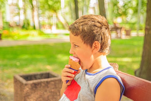 cute young boy in a gray T-shirt sits on a bench in the park on a sunny day and eat an ice cream cone