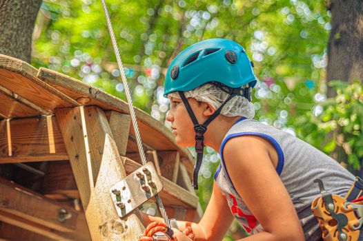profile view of a teenage boy wearing a blue helmet and climbing gear on a ladder in a forest rope adventure park