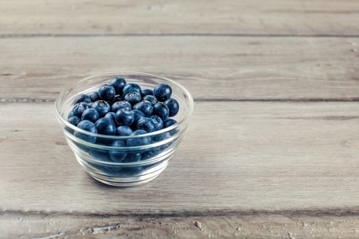 Fresh blueberries in small transparent glass bowl placed on wood desk.