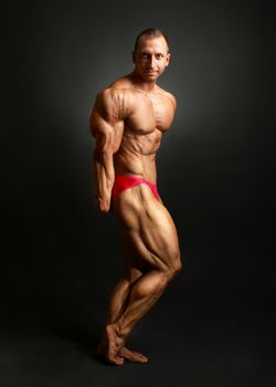 Studio shot of male bodybuilder posing, showing front muscles, huge triceps and legs, with black background.