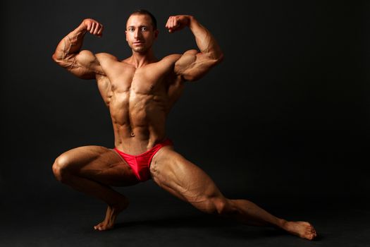 Young male bodybuilder standing knee bend, one leg squat, showing front double biceps pose. Studio photo with black background.