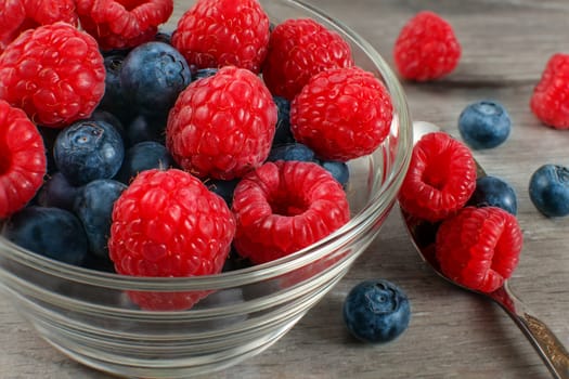 Small glass bowl of fresh raspberries and blueberries with spoon next to it, placed on gray wooden table.