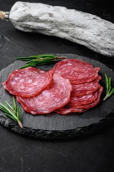 Dry cured, whole and slices cuts of salchichon sausage on black textured surface.
