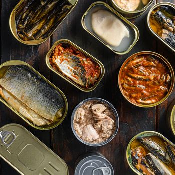 Cans of canned with different types of fish and seafood, over rustic wooden dark planks Top view square.